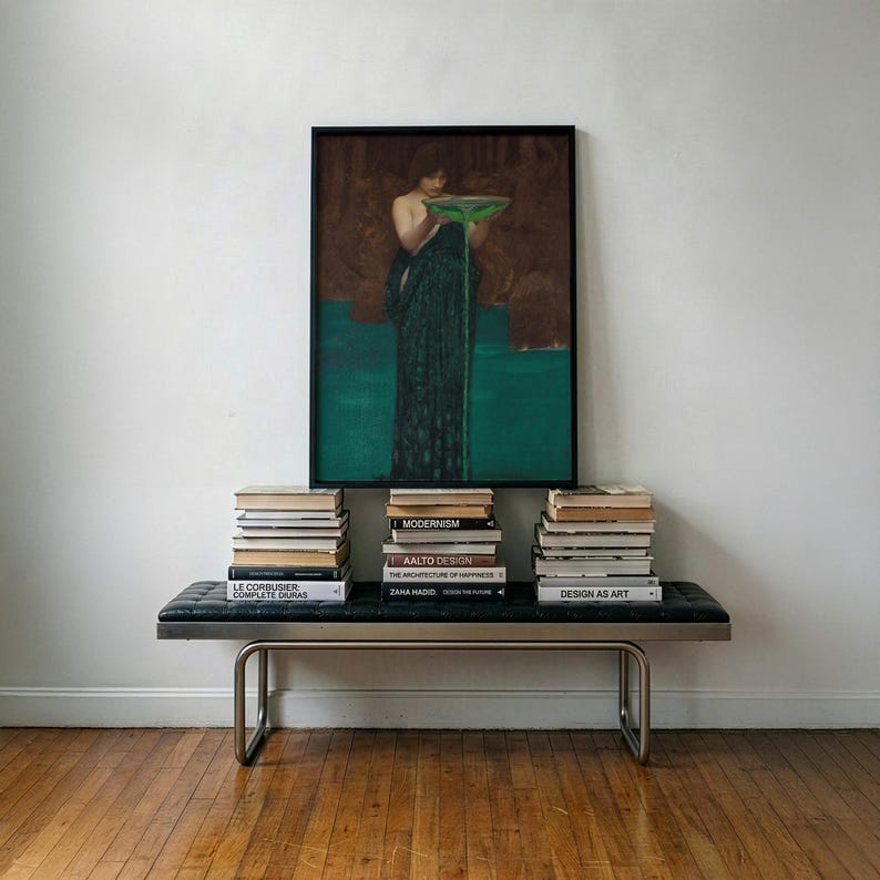 May include: A framed rectangular wall art print depicting a figure in a dark green robe holding a bowl with green liquid pouring out, positioned above a black leather bench with metal legs and three stacks of books including titles like Aalto Design.