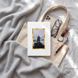 May include: A white notebook featuring a gold-framed print of Caspar David Friedrich's Wanderer above the Sea of Fog, placed inside a beige canvas tote bag with a smartphone, brown-rimmed glasses, a brown leather watch, and a fountain pen.