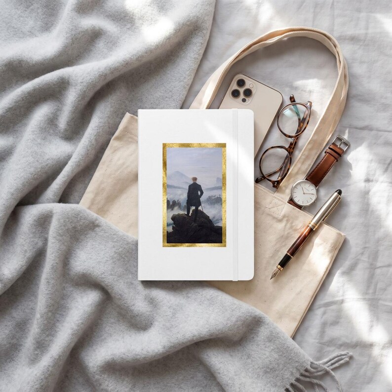 May include: A white notebook featuring a gold-framed print of Caspar David Friedrich's Wanderer above the Sea of Fog, placed inside a beige canvas tote bag with a smartphone, brown-rimmed glasses, a brown leather watch, and a fountain pen.