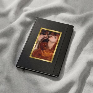 May include: A black hardcover notebook featuring a rectangular print of a painting on the front cover. The artwork depicts a person with reddish hair wearing an orange garment, holding a metallic object near their face, framed in gold.