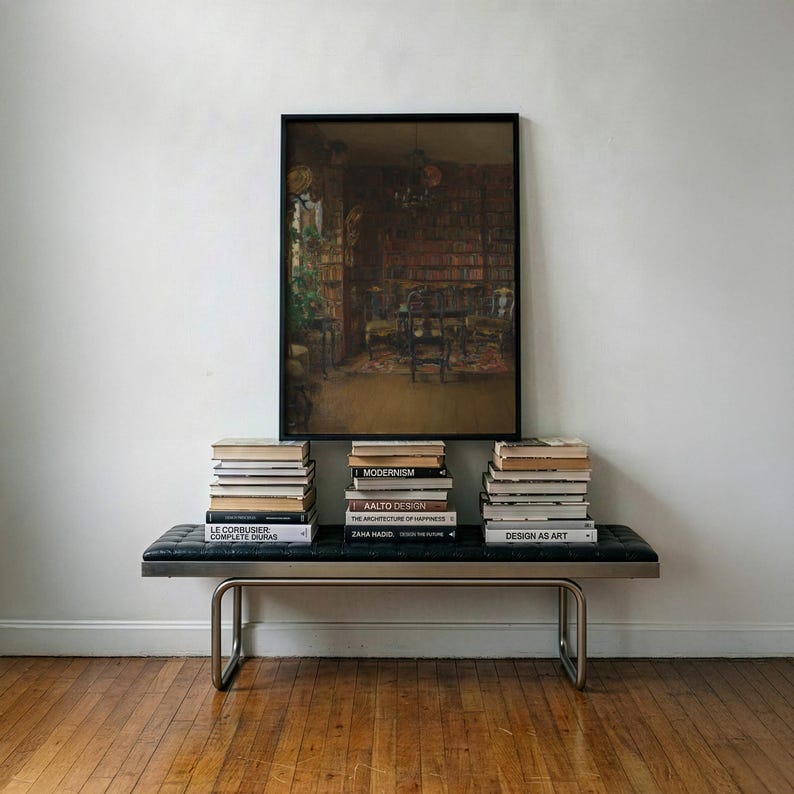 May include: A rectangular framed painting depicting a dark, detailed library interior with bookshelves and furniture, centered above a black tufted leather bench with metal legs, flanked by three stacks of various hardcover books.