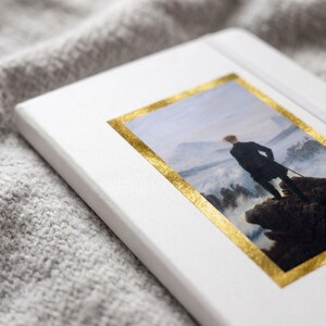 May include: A white notebook featuring a rectangular print of the painting Wanderer above the Sea of Fog by Caspar David Friedrich, framed by a metallic gold border on the front cover, with a smooth, clean surface and rounded corners.