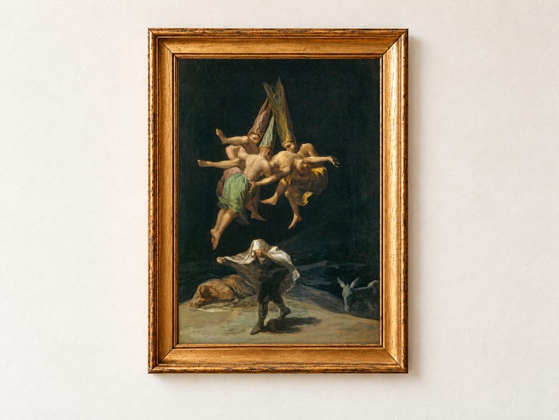 Witches' Flight by Francisco Goya, 1798, as a framed print in an antique gold frame on a cream wall. Three witches in tall pointed hats carry a struggling figure up into a black night sky, while a man below hides under a white sheet beside a donkey.
