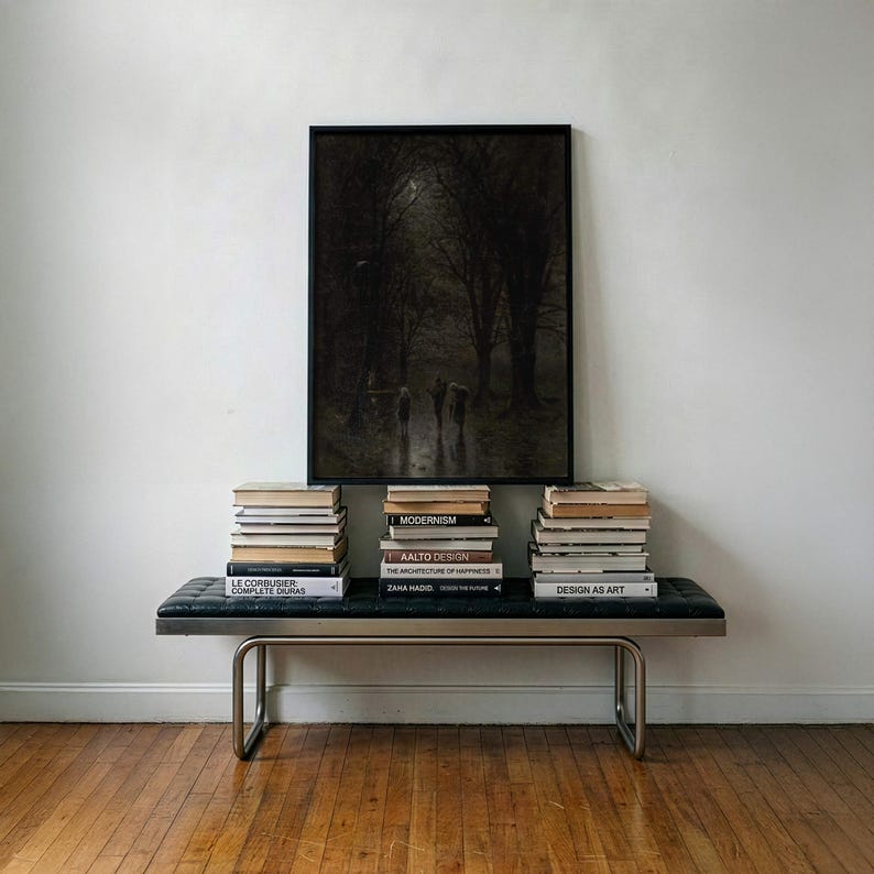 May include: A rectangular black-framed wall art piece depicting a dark, moody forest scene with figures, centered above a black tufted leather bench with a metal frame. Three stacks of books are arranged on the bench, featuring titles like Modernism, Aalto Design, and Le Corbusier.