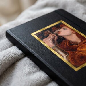 May include: A black hardcover journal featuring a rectangular inset print of a classical painting depicting a person with reddish hair holding a sword. The artwork is framed by a thin gold border, and the journal cover has a textured, matte finish.