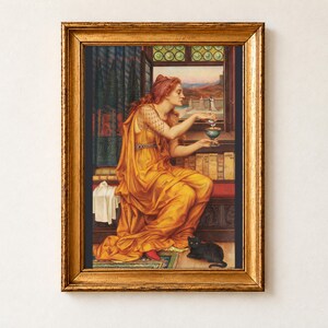 The Love Potion by Evelyn De Morgan, 1903, as a framed print in an antique gold frame on a cream wall. A red-haired sorceress in a golden robe pours a potion into a goblet beside shelves of old books, a black cat with green eyes at her feet.