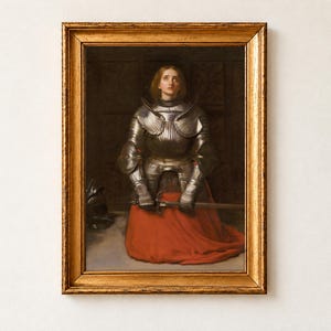 Joan of Arc by John Everett Millais, 1865, as a framed print in an antique gold frame on a cream wall. Joan kneels in prayer in polished steel armour and a red skirt, gazing upward as she holds her sword, her helmet resting on the floor beside her.