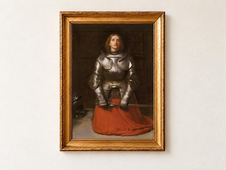 Joan of Arc by John Everett Millais, 1865, as a framed print in an antique gold frame on a cream wall. Joan kneels in prayer in polished steel armour and a red skirt, gazing upward as she holds her sword, her helmet resting on the floor beside her.