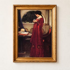 The Crystal Ball by John William Waterhouse, 1902, as a framed print in an antique gold frame on a cream wall. A dark-haired woman in a red velvet gown gazes into a crystal ball, a skull resting on an open book of spells on the table beside her.