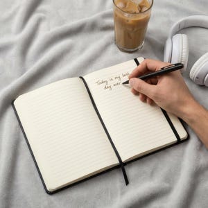 May include: An open notebook with cream-colored lined pages featuring the handwritten text Today is my best day ever. A black pen rests on the page, accompanied by a glass of iced coffee and white over-ear headphones on a soft gray fabric surface.