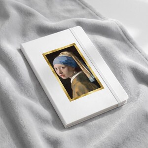 May include: A white notebook with an elastic closure band featuring a rectangular print of the Girl with a Pearl Earring painting. The artwork displays a figure wearing a blue headscarf and a pearl earring, framed by a metallic gold border.