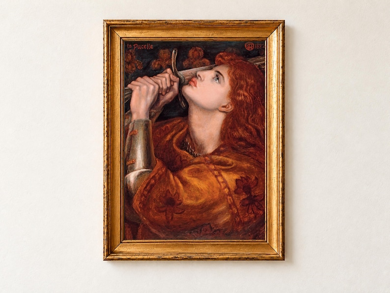Joan of Arc by Dante Gabriel Rossetti, 1882, as a framed print in an antique gold frame on a cream wall. Joan, with long red hair and an orange gown over her armour, kisses the hilt of her sword before a background of red lilies, inscribed La Pucelle.