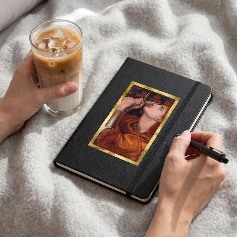 May include: A black hardcover notebook featuring a rectangular print of a painting of a person with red hair holding a sword. The notebook has a black elastic closure and is accompanied by a black ballpoint pen and a glass of iced coffee.