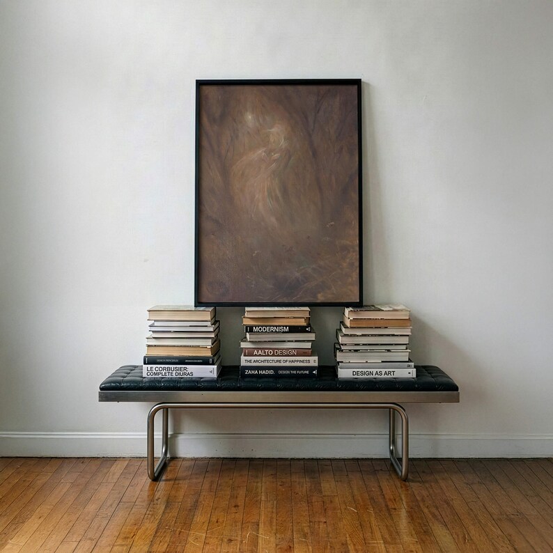 May include: A rectangular framed abstract painting with dark brown and tan tones hangs above a black tufted leather bench with a metal frame. Three stacks of books are arranged on the bench, featuring titles like Modernism and Design as Art.