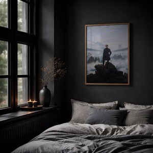 May include: A framed print of Caspar David Friedrich's Wanderer above the Sea of Fog, featuring a person in a dark coat standing on a rocky cliff overlooking a misty mountain landscape, displayed on a dark wall above a bed with gray bedding.
