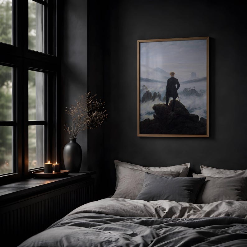 May include: A framed print of Caspar David Friedrich's Wanderer above the Sea of Fog, featuring a person in a dark coat standing on a rocky cliff overlooking a misty mountain landscape, displayed on a dark wall above a bed with gray bedding.