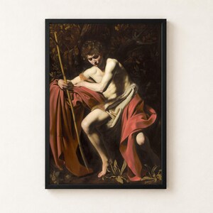 Saint John the Baptist in the Wilderness by Caravaggio, 1604, as a framed print in a black frame on a cream wall. A young man in a scarlet cloak sits in deep shadow holding a thin reed cross, his face turned down in thought, against a dark wilderness.