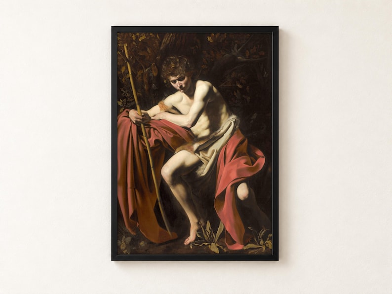 Saint John the Baptist in the Wilderness by Caravaggio, 1604, as a framed print in a black frame on a cream wall. A young man in a scarlet cloak sits in deep shadow holding a thin reed cross, his face turned down in thought, against a dark wilderness.