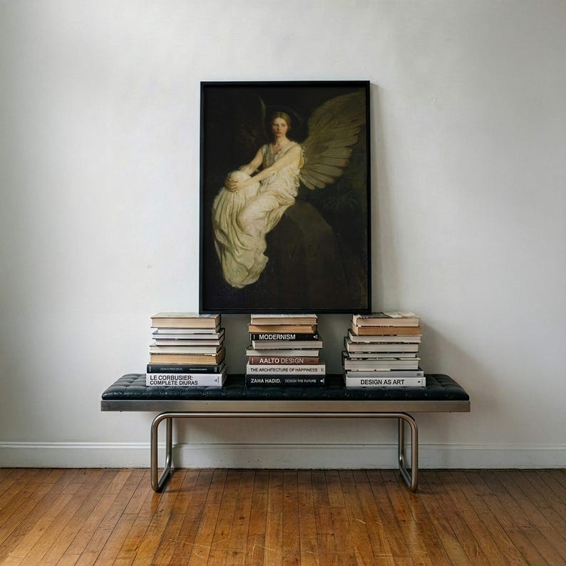 May include: A framed rectangular wall art print depicting a winged figure in a white robe sitting on a dark rock. The artwork is positioned above a black tufted bench with a metal frame, which holds three stacks of various hardcover books.