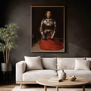 May include: A framed wall art print depicting a person with shoulder-length hair wearing polished steel plate armor over a long, vibrant red skirt. The figure is kneeling, holding a sword horizontally across their lap, with a dark, somber background.