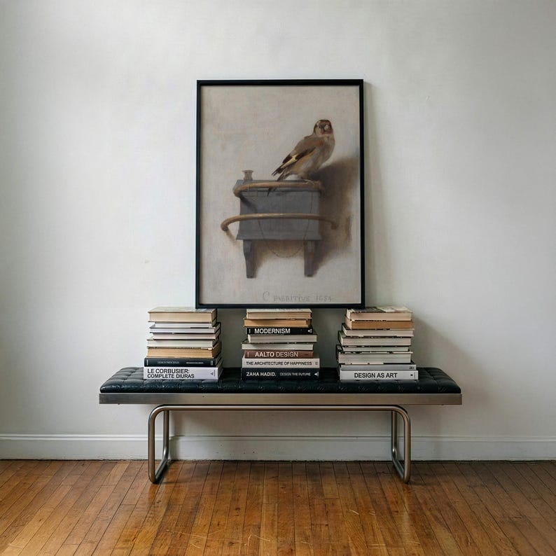 May include: A framed print featuring a brown bird perched on a grey box, displayed above a black leather bench with metal legs. Three stacks of books are arranged on the bench, including titles like Modernism, Aalto Design, and Zaha Hadid.