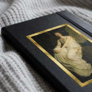 May include: A black notebook with a rectangular gold-bordered inset featuring a classical painting of a winged figure in a white gown. The cover has a textured surface and a black elastic band closure running vertically along the right side.