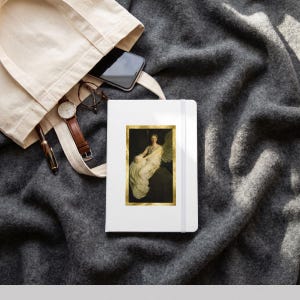 May include: A white notebook featuring a rectangular print of a winged figure in a gold frame, placed next to a beige canvas tote bag containing a black smartphone, a pair of round glasses, and a black pen with a gold clip.