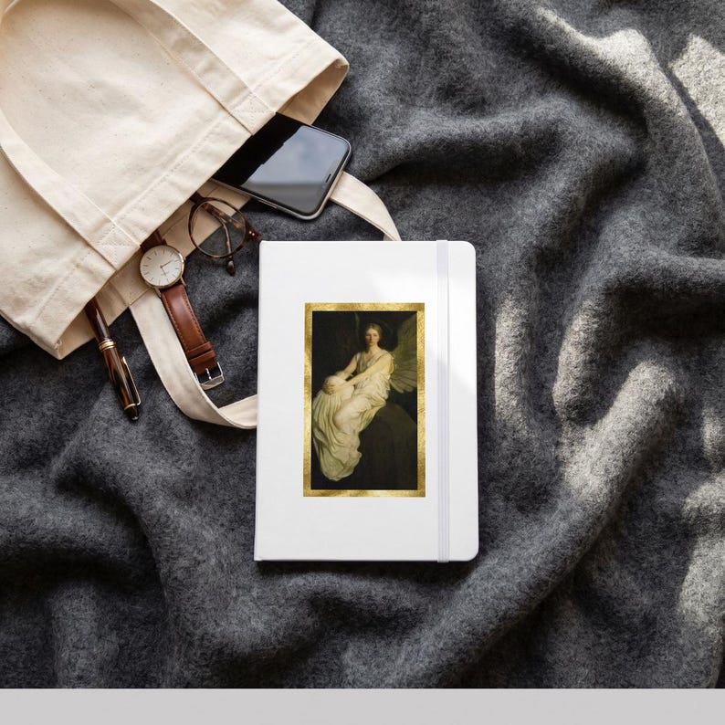 May include: A white notebook featuring a rectangular print of a winged figure in a gold frame, placed next to a beige canvas tote bag containing a black smartphone, a pair of round glasses, and a black pen with a gold clip.