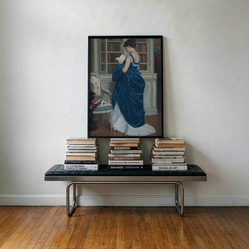 Pu&ograve; includere: Una stampa incorniciata di un dipinto che raffigura una figura in un abito di velluto blu intenso che legge un libro in una biblioteca, sopra una panca in pelle nera trapuntata con telaio in metallo e tre pile di libri come Modernism.
