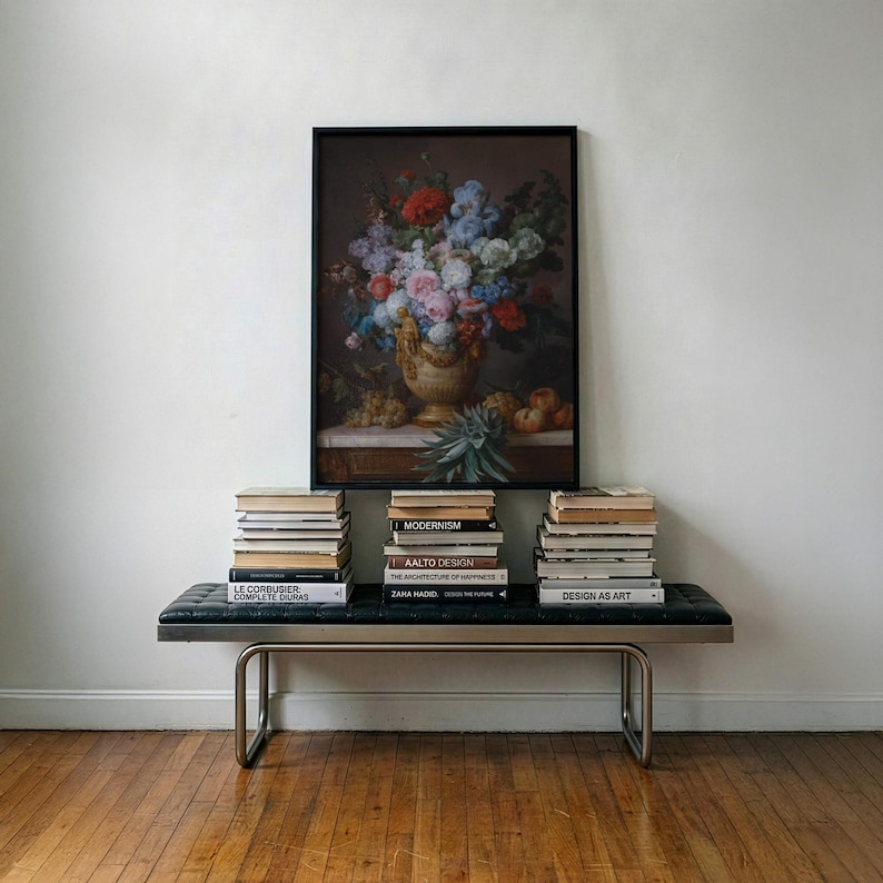 May include: A framed still life painting of a floral arrangement in a decorative vase with fruit, displayed above a black tufted bench with metal legs. Three stacks of books are arranged on the bench, featuring titles like Modernism, Aalto Design, and Design as Art.
