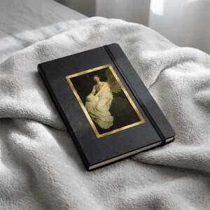 May include: A black hardcover notebook featuring a rectangular gold-framed print of a classical painting depicting a seated figure in a white gown with wings. The notebook includes a vertical black elastic band closure attached to the front cover.