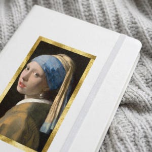 May include: A white notebook featuring a rectangular print of the painting Girl with a Pearl Earring, framed by a gold border. The notebook has a white elastic closure band attached to the side, providing a secure way to keep the cover closed.