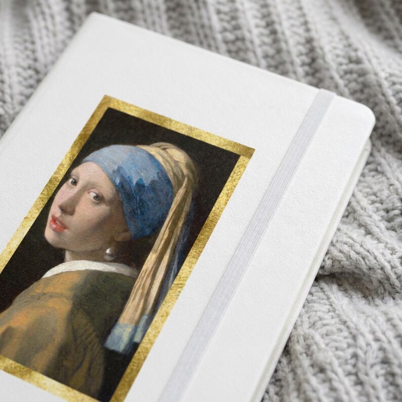 May include: A white notebook featuring a rectangular print of the painting Girl with a Pearl Earring, framed by a gold border. The notebook has a white elastic closure band attached to the side, providing a secure way to keep the cover closed.