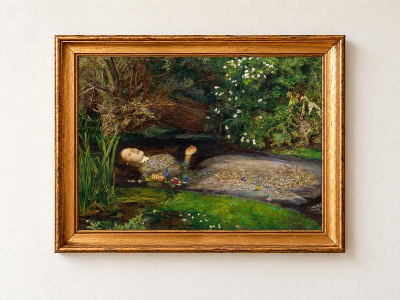 Ophelia by John Everett Millais, 1851 to 1852, as a framed print in an antique gold frame on a cream wall. Ophelia floats on her back in a quiet river, her silver embroidered dress spread on the water and loose flowers drifting from her hand beneath overhanging willows and wild roses.