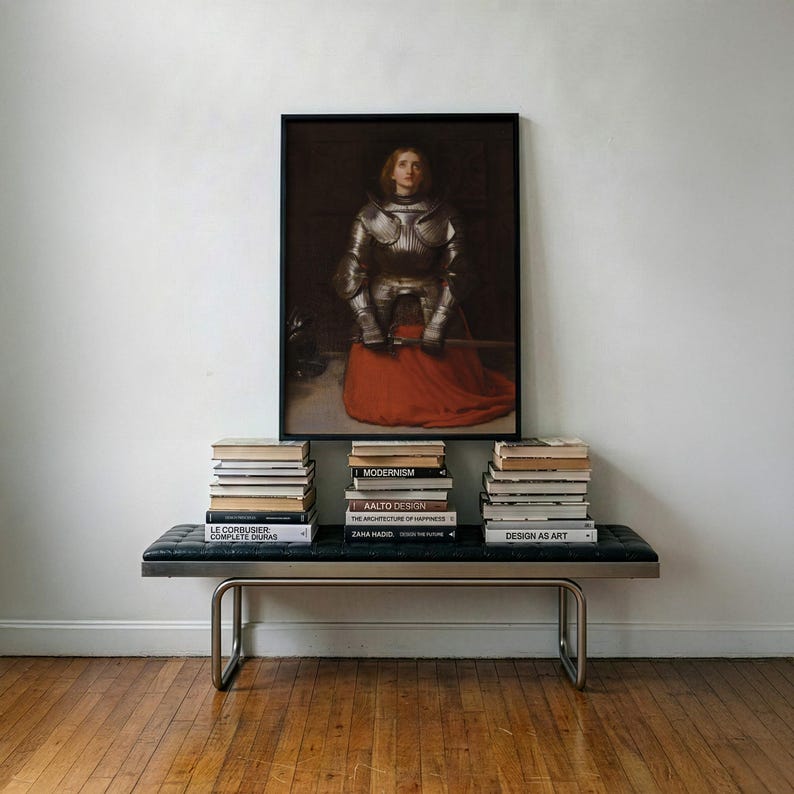 May include: A framed painting of a figure in silver plate armor wearing a red skirt, positioned above a black tufted bench with a metal frame. Three stacks of books are arranged on the bench, featuring titles like Modernism and Zaha Hadid.
