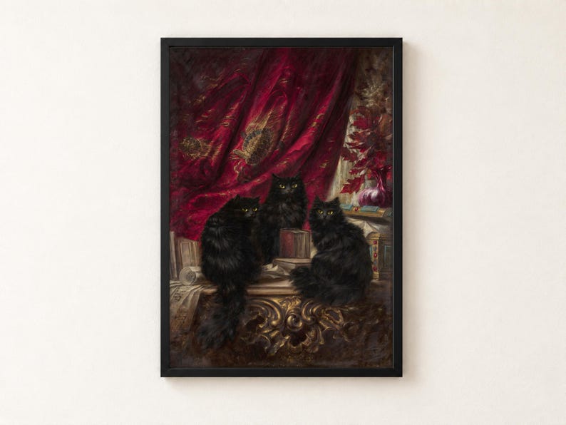 Three Black Cats by Carl Kahler as a framed print in a black frame on a cream wall. Three long-haired black cats with yellow eyes sit on a gilded table among books and scrolls, in front of a crimson velvet curtain embroidered in gold.