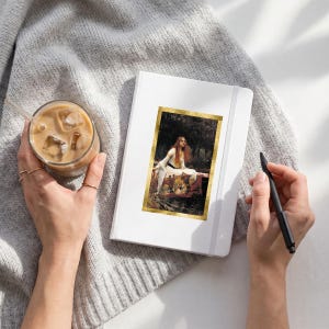 May include: A white hardcover notebook featuring a rectangular print of John William Waterhouse's The Lady of Shalott painting with a gold border. The notebook is placed on a gray knit fabric next to a glass of iced coffee and a black pen.