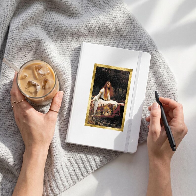 May include: A white hardcover notebook featuring a rectangular print of John William Waterhouse's The Lady of Shalott painting with a gold border. The notebook is placed on a gray knit fabric next to a glass of iced coffee and a black pen.