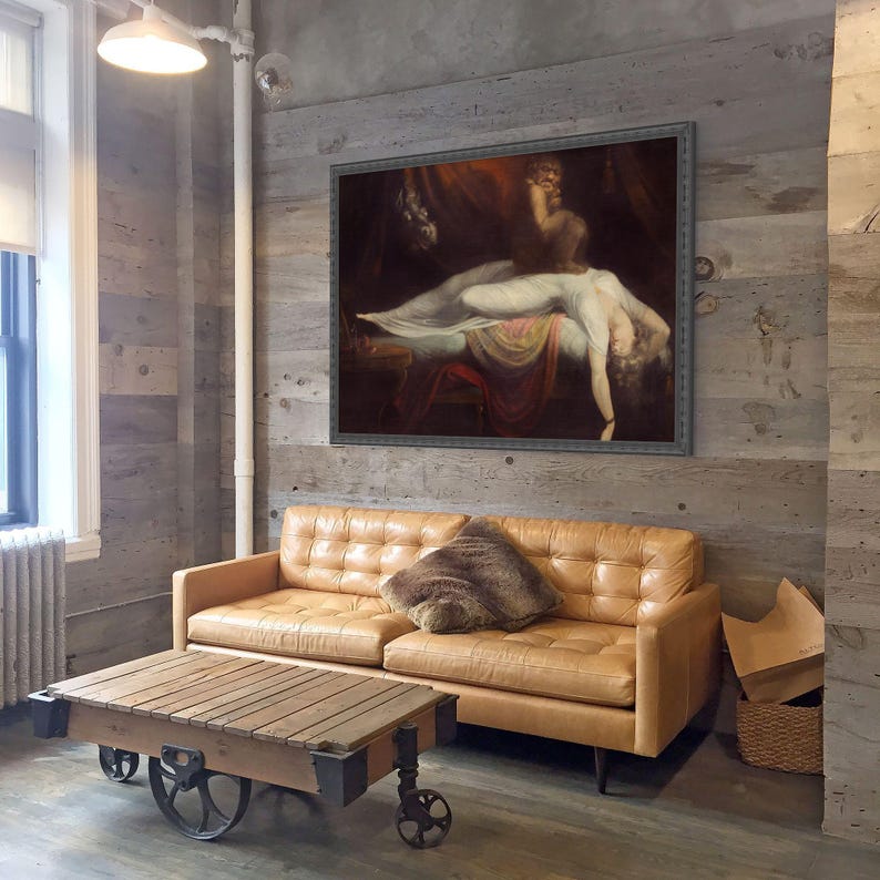 May include: A framed oil painting depicting a reclining figure in a white garment with a dark, shadowy creature perched above, set against a dark background, displayed in a room with a tan leather sofa and a wooden cart coffee table.