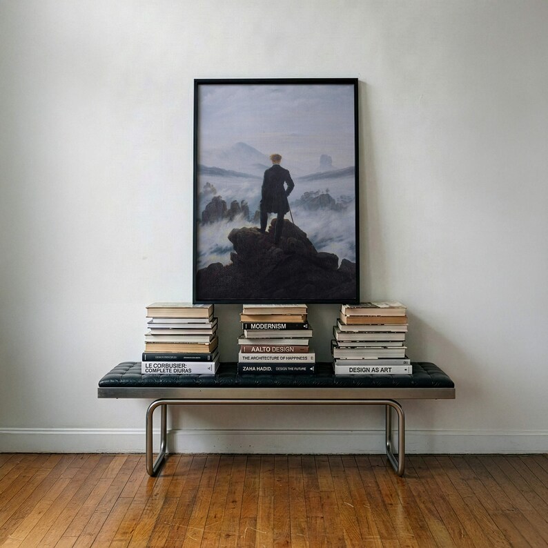 May include: A framed print of Wanderer above the Sea of Fog by Caspar David Friedrich, featuring a person in a dark coat standing on a rocky cliff overlooking a misty mountain landscape, displayed above a black leather bench with metal legs.
