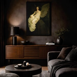 May include: A framed wall art print depicting a winged figure in a white robe sitting on a stone ledge. The artwork is displayed above a dark wood sideboard, a black table lamp, a black bowl, and a stack of books in a dimly lit room.