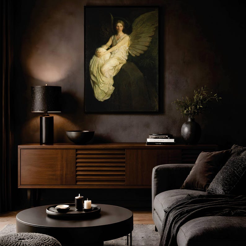 May include: A framed wall art print depicting a winged figure in a white robe sitting on a stone ledge. The artwork is displayed above a dark wood sideboard, a black table lamp, a black bowl, and a stack of books in a dimly lit room.