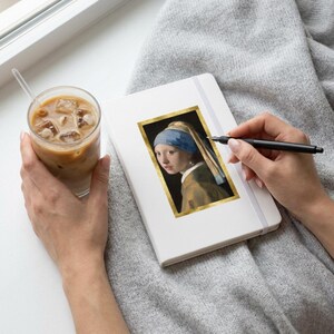 May include: A white notebook featuring a rectangular sticker of the Girl with a Pearl Earring painting with a gold border. The notebook is held by hands, with one hand holding a black pen and the other holding a glass of iced coffee.