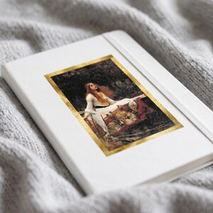 May include: A white hardcover notebook featuring a rectangular print of a painting depicting a person with long red hair in a white dress, framed by a metallic gold border. The notebook has a white elastic closure band attached to the cover.