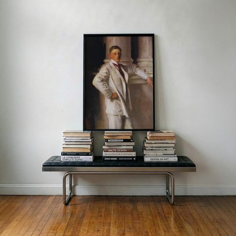 May include: A framed portrait painting of a person in a white suit with a red tie, displayed above a black tufted bench with a metal frame. Three stacks of books are arranged on the bench, featuring titles like Modernism and Zaha Hadid.