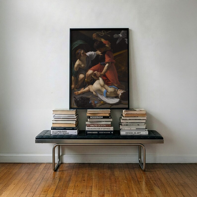 May include: A rectangular framed print of a classical painting featuring figures in red and white robes, positioned above a black tufted leather bench with a metal frame. Three stacks of books are arranged on the bench beneath the artwork.