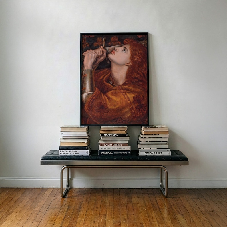 May include: A framed rectangular print featuring a painting of a person with long red hair holding a sword near their face, wearing a golden garment. The artwork is displayed above a black leather bench with three stacks of books.