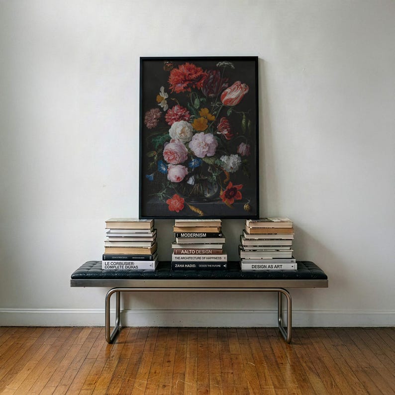 Puede incluir: Una l&aacute;mina rectangular enmarcada que muestra un bodeg&oacute;n floral oscuro con flores rojas, rosas, blancas y naranjas en un jarr&oacute;n. La l&aacute;mina descansa sobre un banco de cuero negro con estructura met&aacute;lica, flanqueado por dos pilas de libros.