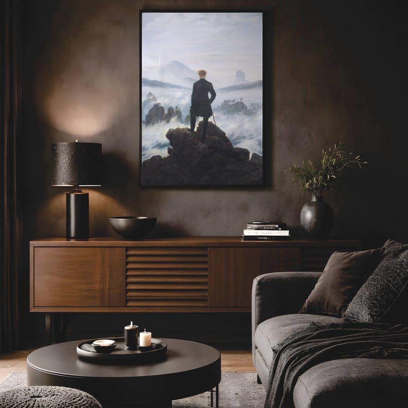 May include: A framed print of Caspar David Friedrich's Wanderer above the Sea of Fog, depicting a person in a dark coat standing on a rocky cliff overlooking a misty mountain landscape, displayed on a dark wall above a wooden sideboard.