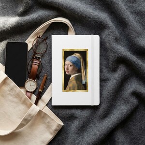 May include: A white notebook featuring a rectangular print of the Girl with a Pearl Earring painting framed in gold. The notebook is placed next to a black smartphone, a brown leather-strap wristwatch, and a brown pen inside a beige tote bag.